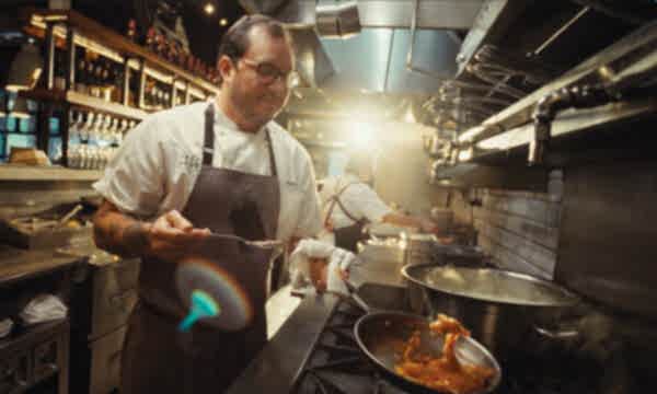 A person in a professional kitchen cooks with focus, surrounded by cookware and shelves of bottles, creating a vibrant culinary scene.