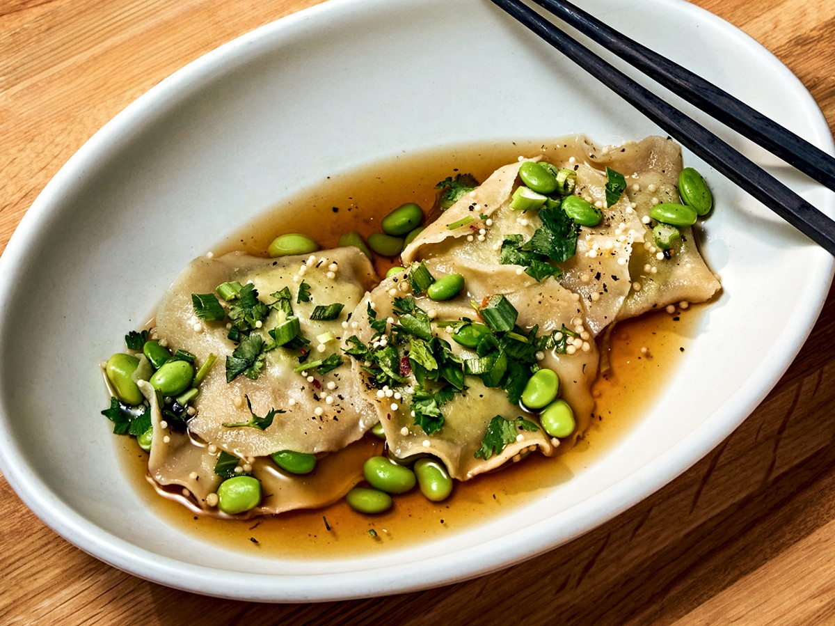 A plate of dumplings garnished with herbs and edamame, served in soy-based sauce, accompanied by chopsticks on a wooden table.