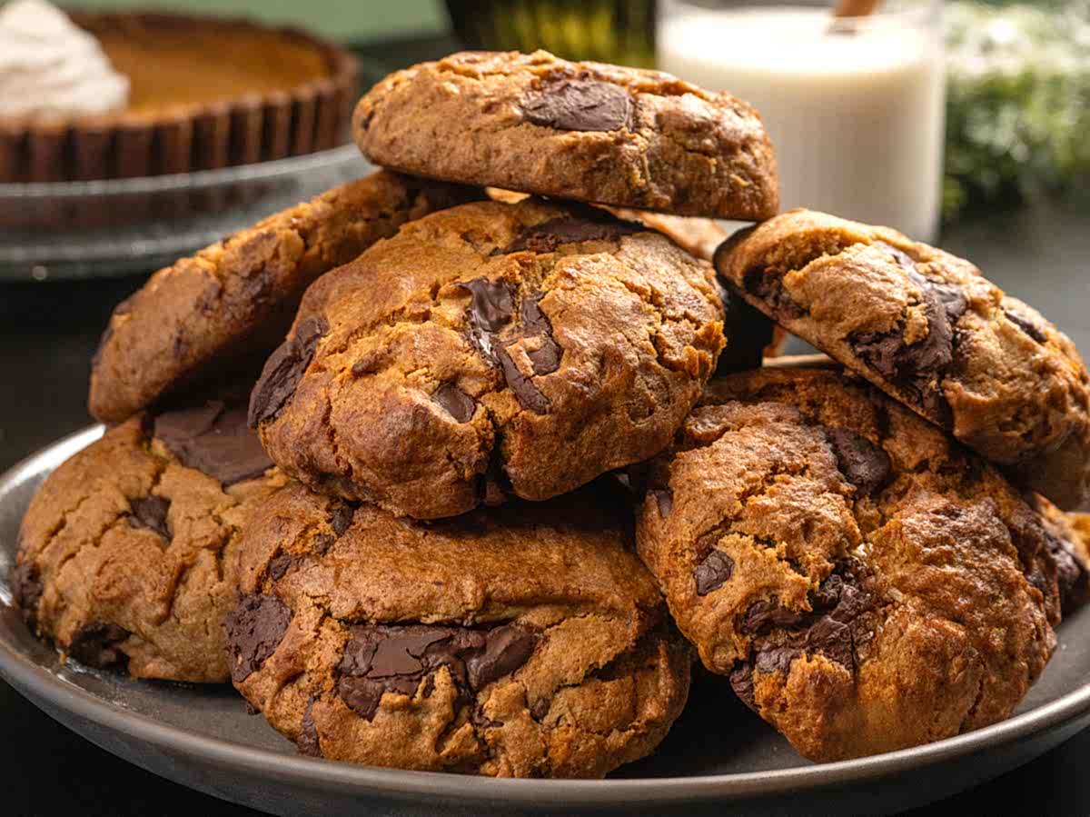A stack of chunky chocolate chip cookies on a plate, with a glass of milk and a pie in the background.