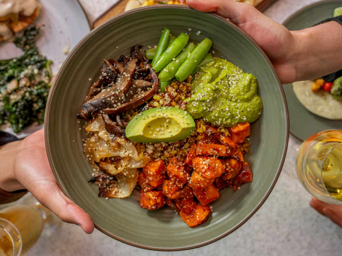 A person holds a bowl of quinoa, avocado, mushrooms, green beans, and sweet potatoes. Another person holds a glass of white wine.