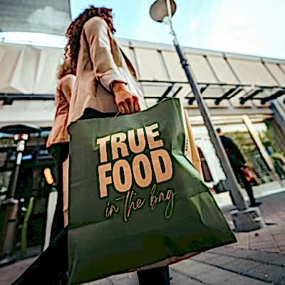 A person carries a green shopping bag reading "TRUE FOOD in the bag" while walking on a street lined with shops.