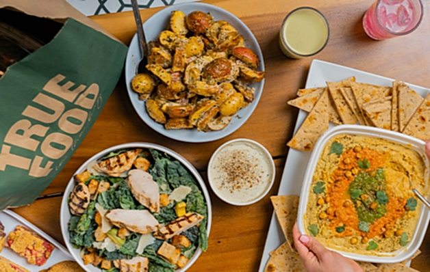 A wooden table filled with vibrant dishes including salad, hummus, roasted potatoes, and drinks, next to a "True Food" bag and pita bread.