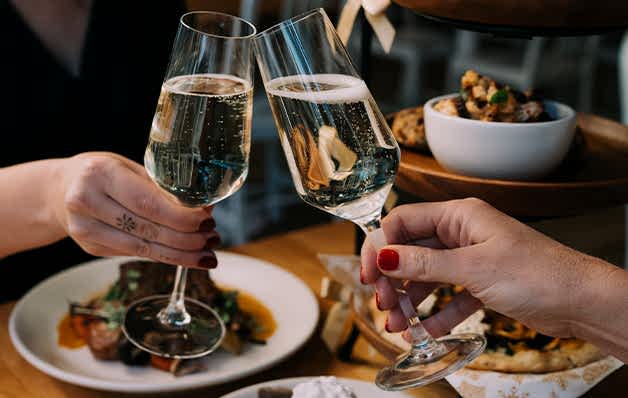 Two people clink champagne glasses over a wooden table with plates of gourmet food, showcasing a celebratory dining experience.