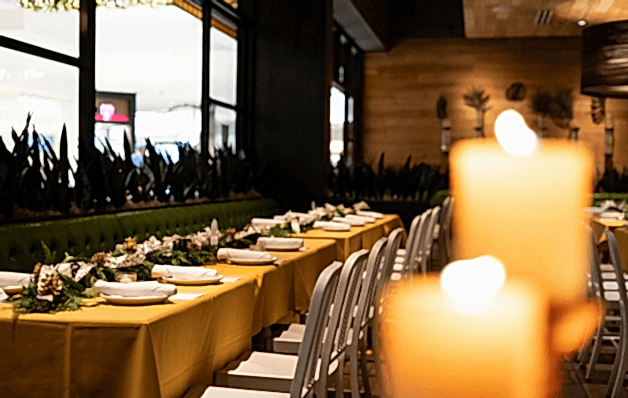 A warmly lit restaurant features tables set with yellow cloths, white chairs, and decorative garlands, with blurred candles in the foreground.