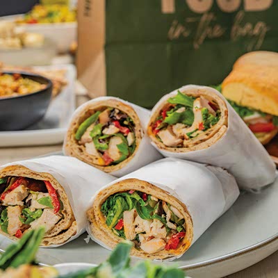A plate with four wrapped chicken and vegetable wraps. A sandwich and salad bowl appear in the blurry background on a table.