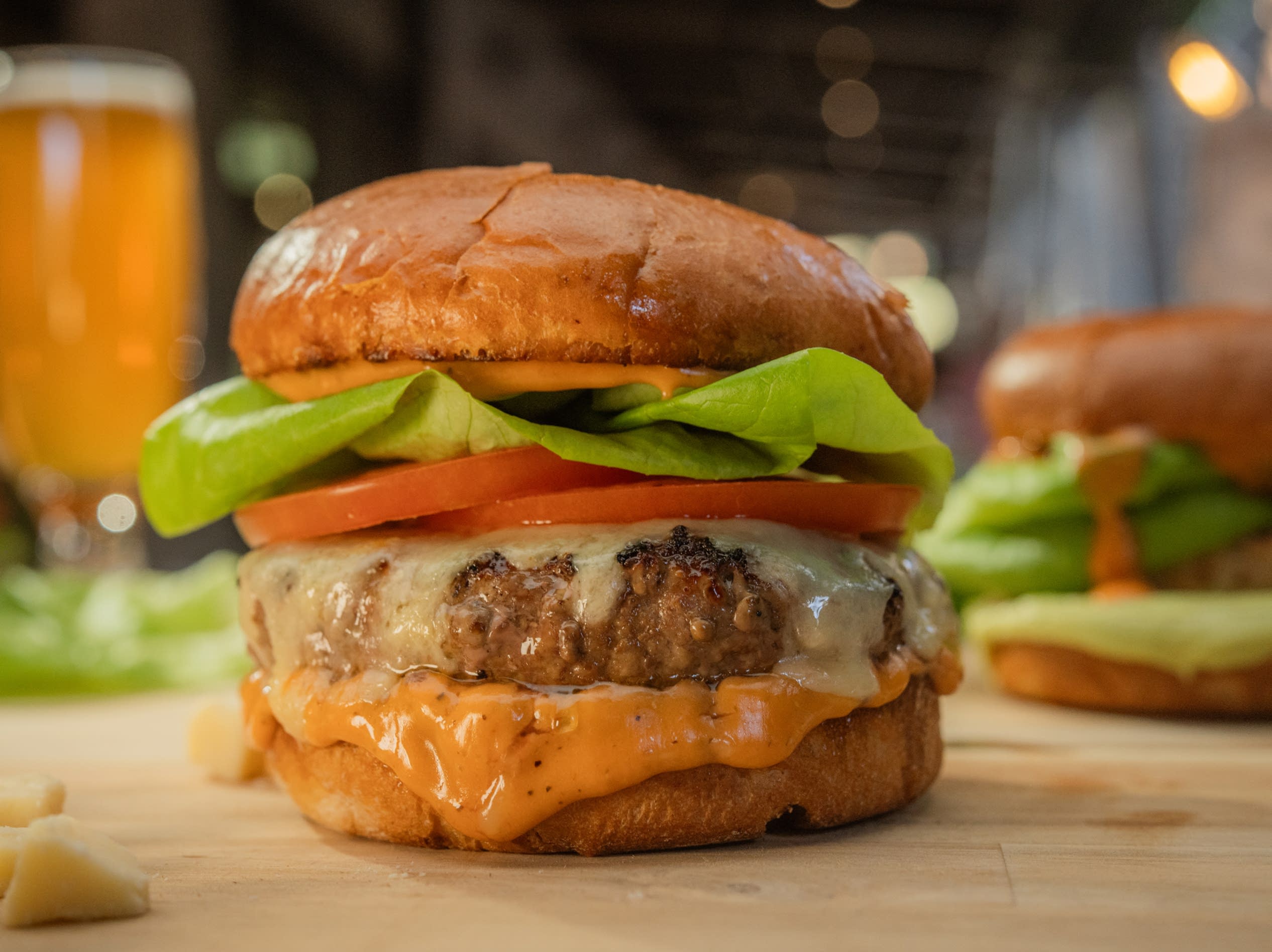 A juicy cheeseburger with lettuce and tomato sits on a wooden surface. A drink is blurred in the background.