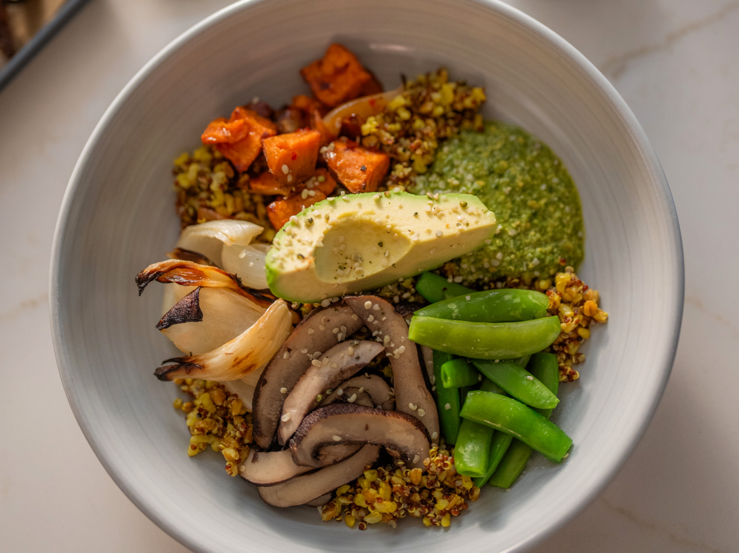 A bowl with roasted sweet potatoes, avocado, mushrooms, sugar snap peas, grilled onions, and green sauce, garnished with seeds.