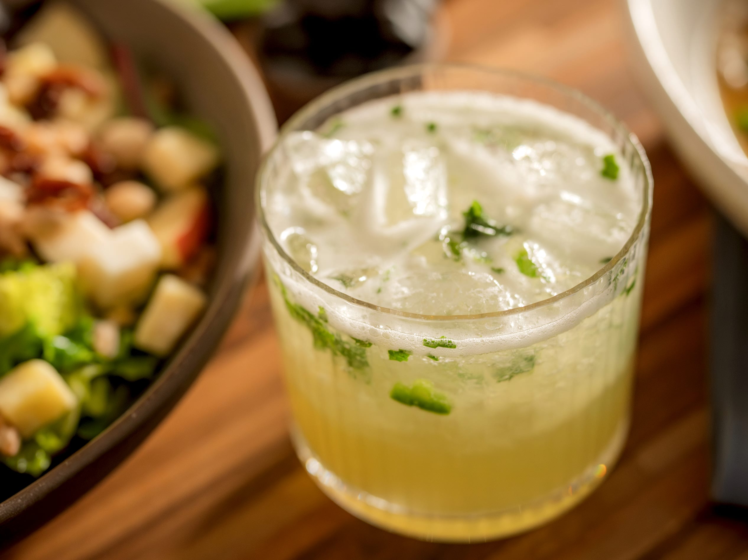 A glass of iced beverage with mint, beside a fresh salad in a bowl, placed on a wooden surface.