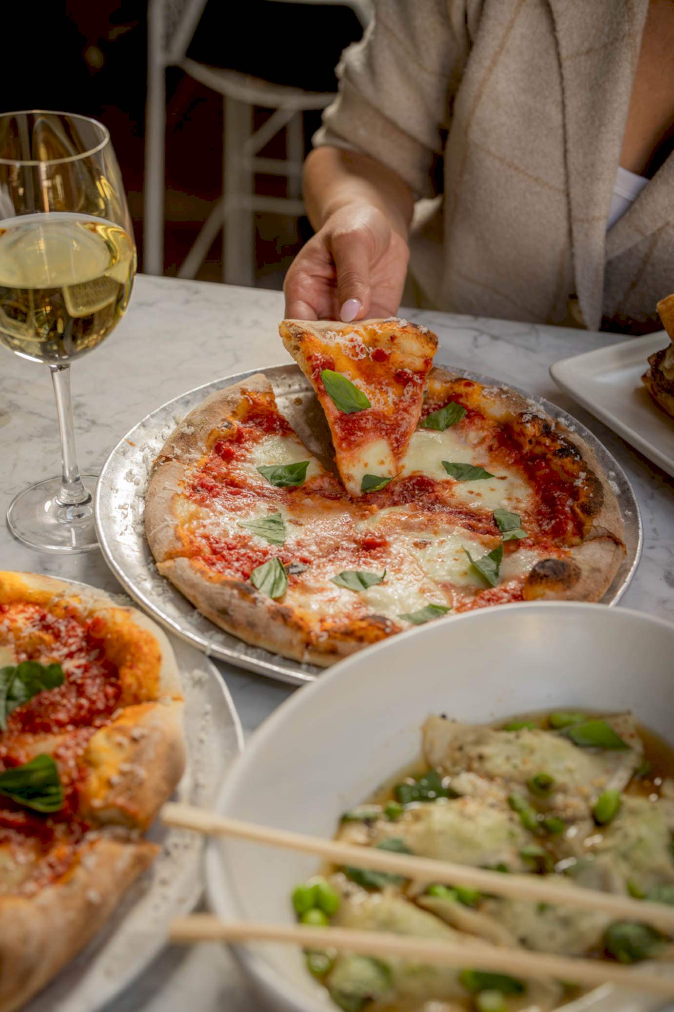 A person enjoys Margherita pizza and dumplings at a restaurant table, accompanied by a glass of white wine.