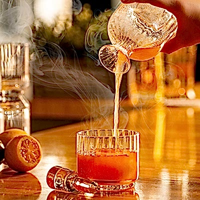 A person pours a steaming beverage into a glass on a wooden bar. Lemons and a small dropper bottle are nearby.